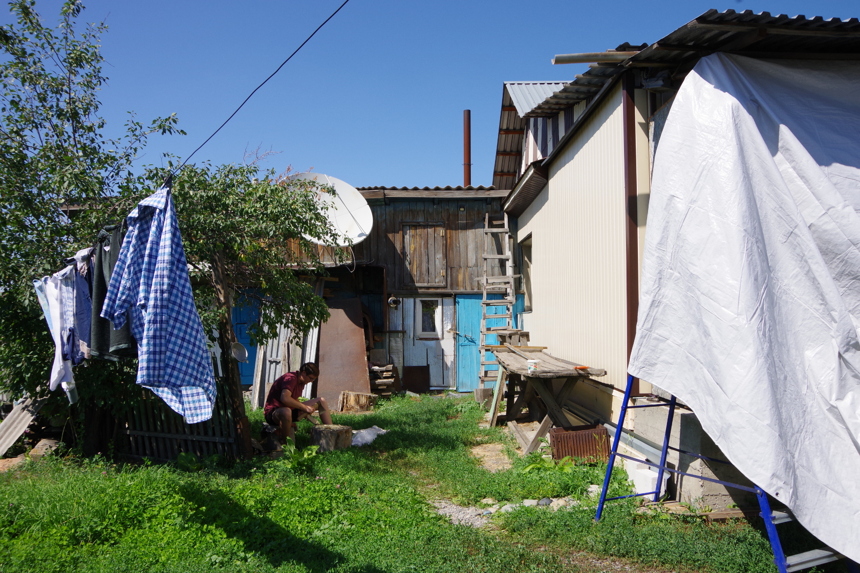 Reinhard beim Schnitzen, dahinter links die Banja (= Sauna), dahinter rechts der Heizungsraum, rechts neben ihm das Haus